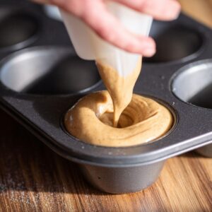 Close-up of a hand piping creamy coffee cake donut batter from a piping bag into a nonstick donut pan on a dark wood countertop. Natural, warm kitchen lighting highlights the smooth texture of the batter, ideal for step-by-step baking tutorials, recipe blogs, or Pinterest posts.