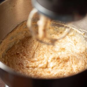 Close-up of creamy coffee cake donut batter with cinnamon specks being mixed in a metal stand mixer bowl, ready to pour into a donut pan. Warm kitchen lighting highlights the texture and color, ideal for step-by-step baking tutorials, recipe blogs, or Pinterest posts