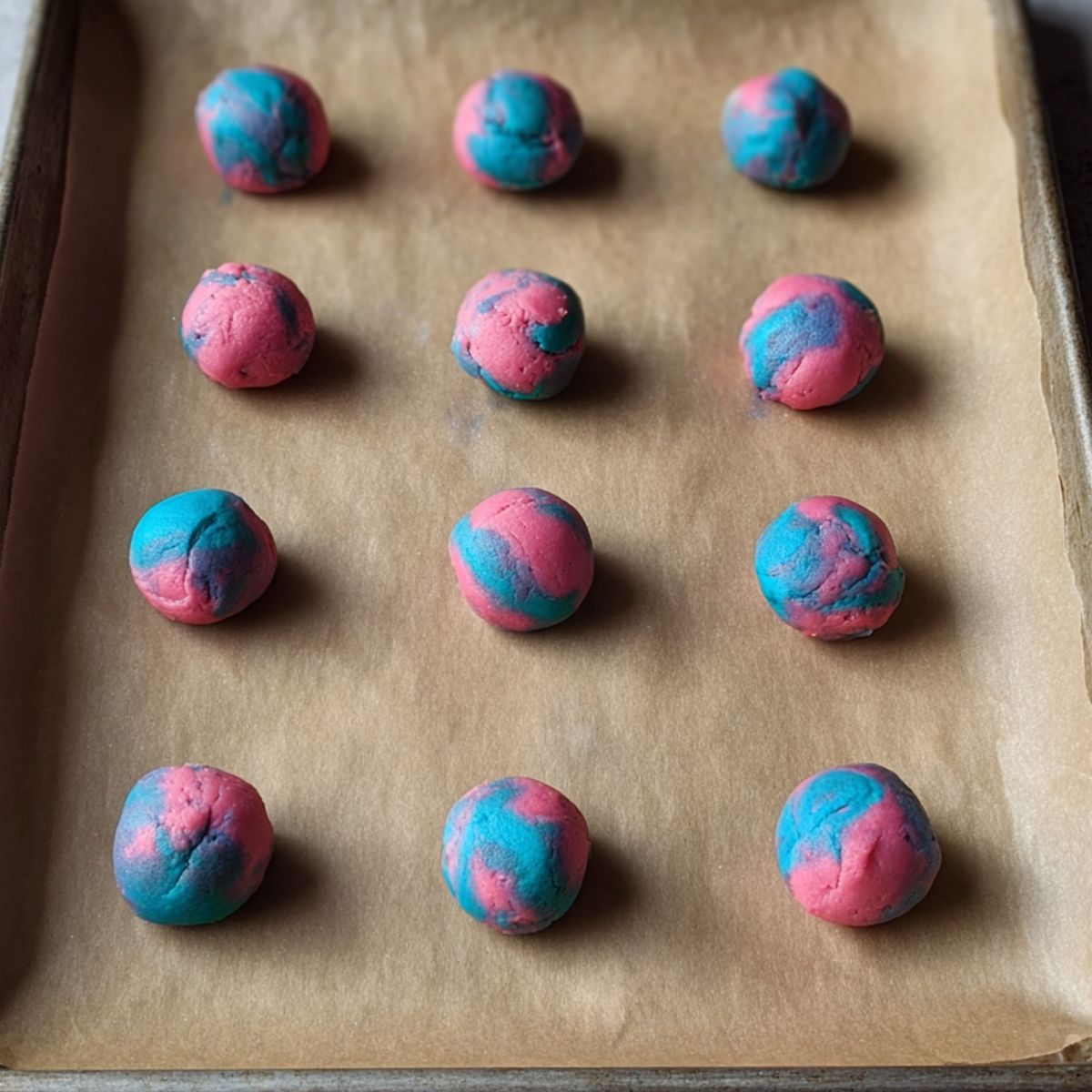 Easy Cotton Candy Cookies Recipe 12 Baking sheet lined with parchment paper holding several small balls of marbled pink and blue cookie dough, spaced evenly for baking.