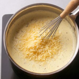 The Best Creamy Salmon Pasta Recipe 16 "Grated cheese being whisked into a creamy sauce in a saucepan."