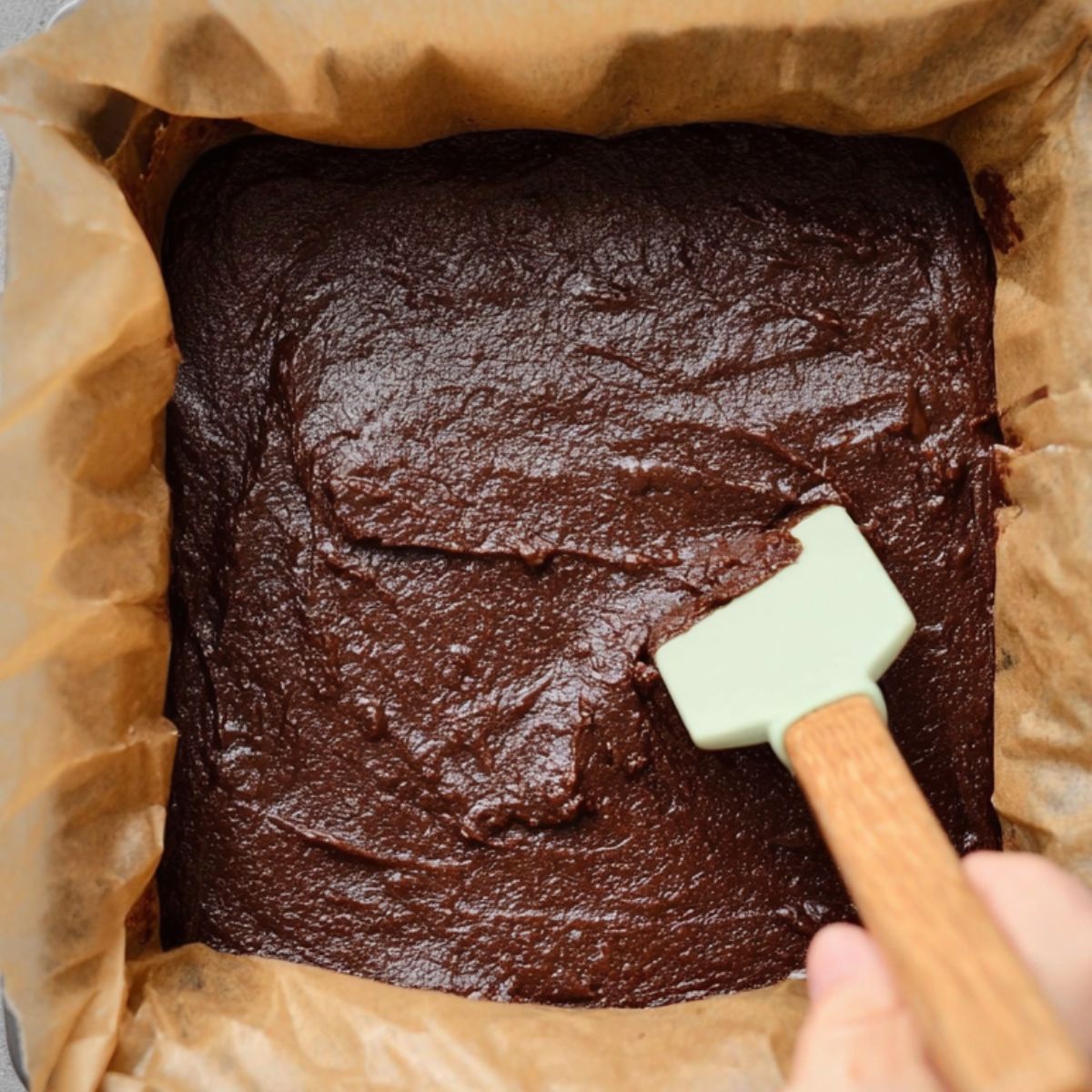 The Best Date Brownies Recipe 13 Chocolate brownie batter being spread in a parchment-lined baking pan using a silicone spatula.