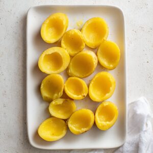 Overhead view of small yellow potato halves with the centers scooped out, arranged on a white plate, ready to be filled with deviled potato mixture.