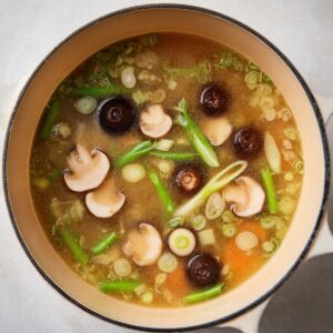 Best Ginger Miso Brothy Soup Recipe 17 A Dutch oven filled with light-colored miso-ginger broth. Sliced shiitake mushrooms, thinly sliced green onions, and other small vegetables float on the surface. The soup is clear, warm, and inviting.