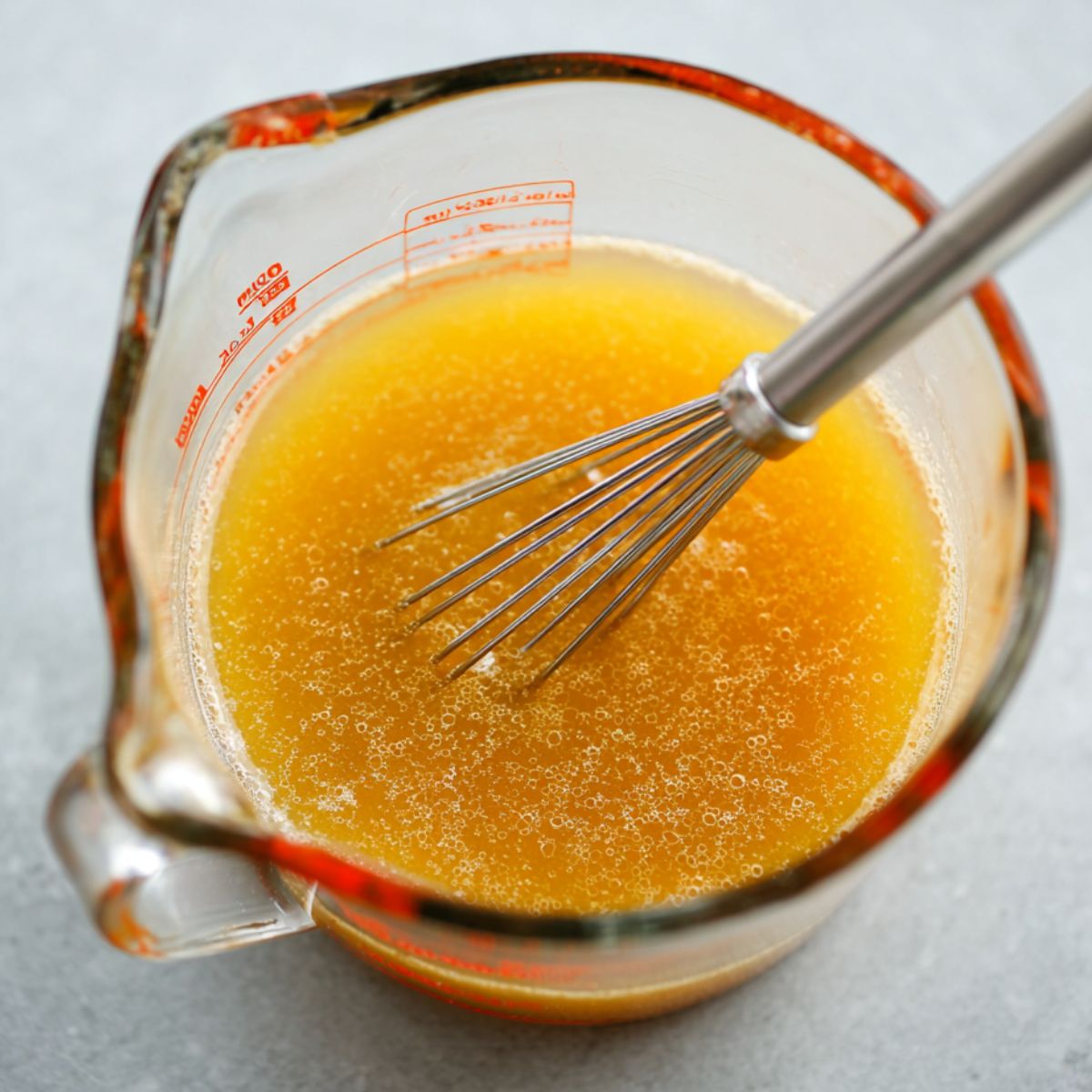 Best Ginger Miso Brothy Soup Recipe 12 A clear glass measuring cup containing a golden-orange broth mixture, with a small metal whisk resting inside. The liquid appears smooth with tiny bubbles, likely a ginger-miso mixture being prepared for the soup.