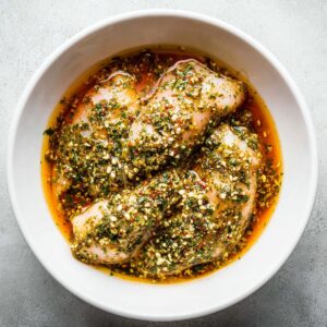 Healthy Greek Marinated Chicken Recipe 15 "Raw chicken breasts coated with a Greek herb and garlic marinade in a white bowl, ready to marinate."