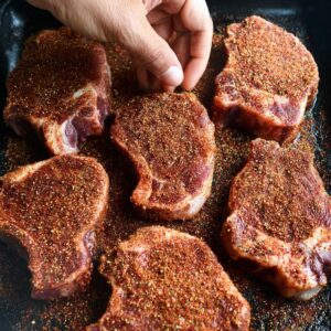 Easy Grilled Spiced Pork Chops Recipe 16 Six raw pork chops evenly coated with a reddish-brown dry rub, pressed gently with a hand to adhere the spices before grilling.