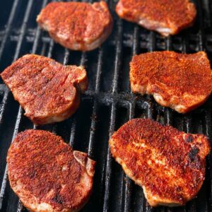 Easy Grilled Spiced Pork Chops Recipe 17 Six pork chops on a grill, each coated in a reddish spice rub, cooking over black grill grates, beginning to sear with some dark grill marks forming.