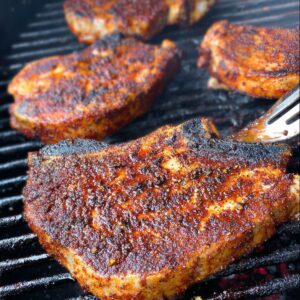 Easy Grilled Spiced Pork Chops Recipe 18 Close-up of four pork chops on a grill, seared with a darkened, caramelized crust, showing the texture of the spice rub and lightly charred edges.