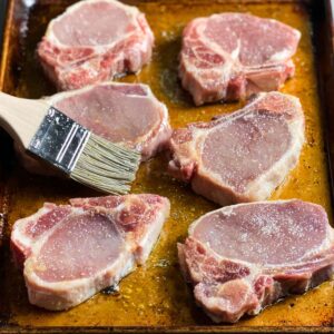 Easy Grilled Spiced Pork Chops Recipe 15 Six raw pork chops on a baking sheet, being brushed with oil or marinade, showing their pink flesh and white marbling, ready for seasoning.