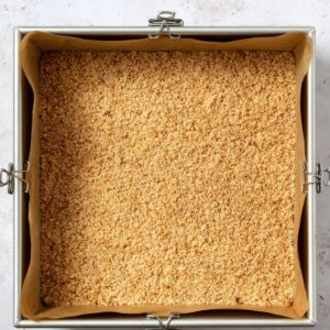 Healthy Key Lime Pie Bars Recipe 16 A square baking pan lined with parchment paper contains a pressed, even layer of crumbly graham cracker crust. The crust is golden brown, and binder clips secure the parchment to the pan edges.