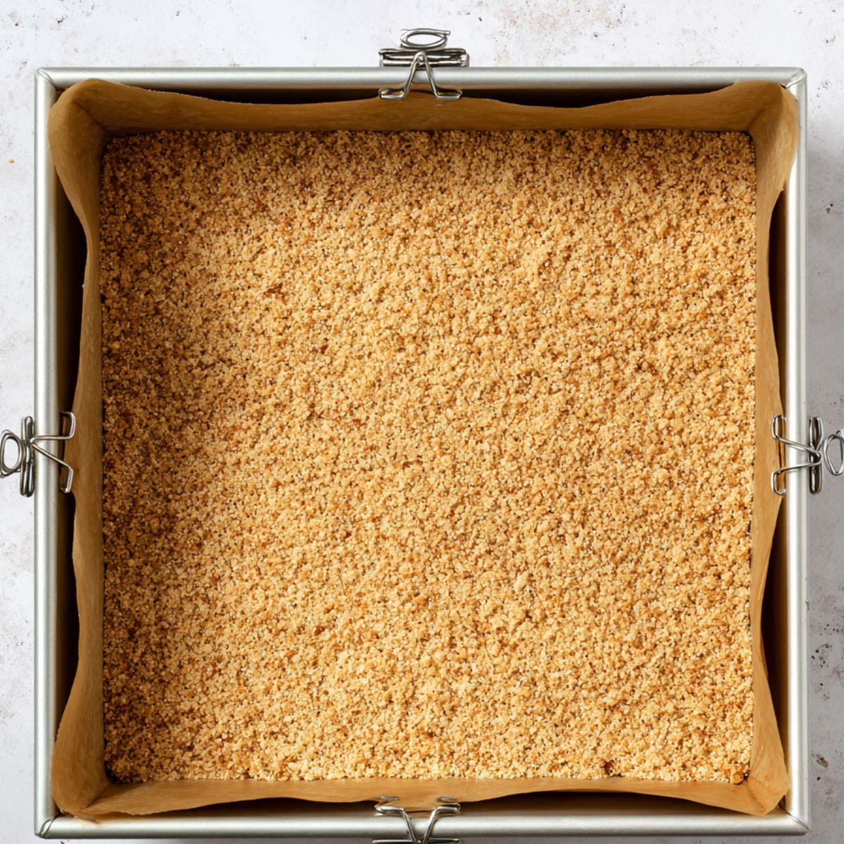 Healthy Key Lime Pie Bars Recipe 11 A square baking pan lined with parchment paper contains a pressed, even layer of crumbly graham cracker crust. The crust is golden brown, and binder clips secure the parchment to the pan edges.