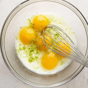 Healthy Key Lime Pie Bars Recipe 17 Four egg yolks in a clear glass mixing bowl with a whisk. The yolks are sprinkled with lime zest, ready to be whisked into the Key Lime Pie filling. The bowl sits on a light, neutral countertop.