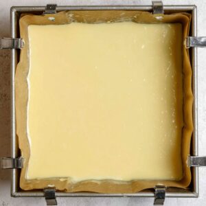 Healthy Key Lime Pie Bars Recipe 18 A square baking pan lined with parchment paper is filled with smooth, pale-yellow Key Lime Pie filling, ready to be baked. The pan is secured with binder clips on the edges to hold the parchment paper in place. The filling surface is glossy and evenly spread.
