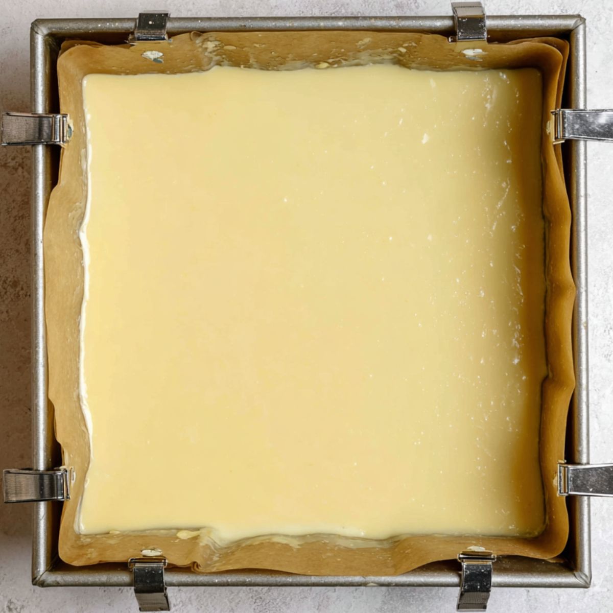 Healthy Key Lime Pie Bars Recipe 13 A square baking pan lined with parchment paper is filled with smooth, pale-yellow Key Lime Pie filling, ready to be baked. The pan is secured with binder clips on the edges to hold the parchment paper in place. The filling surface is glossy and evenly spread.