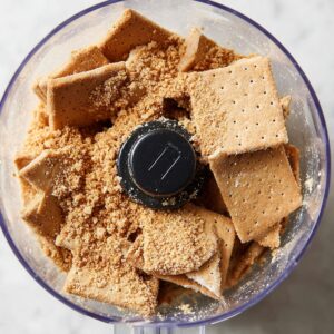 Healthy Key Lime Pie Bars Recipe 15 Several broken graham crackers inside a clear food processor bowl. The crackers are ready to be pulsed into fine crumbs for the pie crust. The background is a light countertop, giving focus to the crackers.