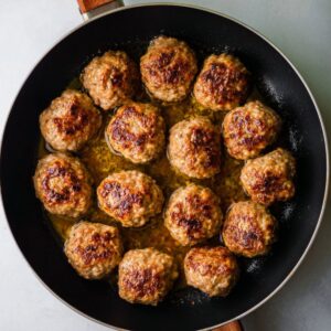 Best Homemade Kotleti Recipe 18 A frying pan filled with golden-brown cooked meatballs, arranged in a circular pattern, sitting in a thin layer of juices.