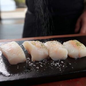 Best Mediterranean Cod Recipe 14 Four raw cod fillets placed on a black cutting board, being lightly sprinkled with salt and seasoning by a person wearing a black apron.