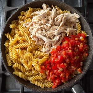 Easy Monterey Chicken Skillet Recipe 14 A skillet with uncooked rotini pasta, shredded chicken, and diced tomatoes, showing ingredients before mixing.