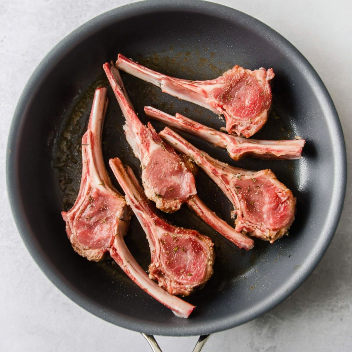 The Best Pan-Seared Lamb Chops Recipe 12 A gray skillet with five raw lamb chops laid flat, showing their pink meat and white bones, ready to be seared.