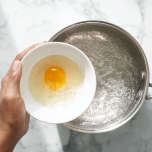 The Best Poached Eggs Recipe 15 Top-down view of a hand holding a small white bowl with a raw egg over a pot of boiling water, preparing to poach the egg, stainless steel pot, white marble countertop, bright natural lighting.