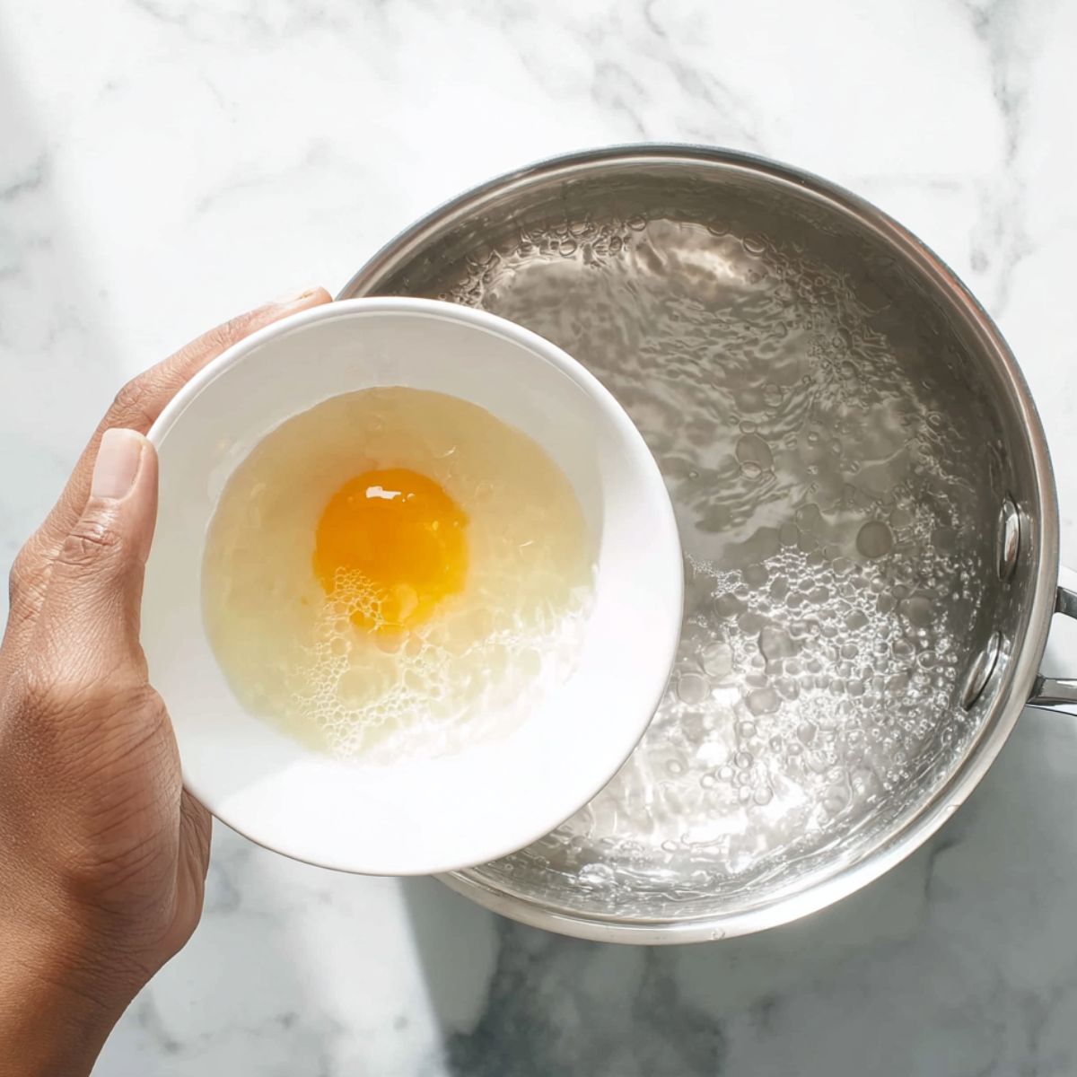 The Best Poached Eggs Recipe 11 Top-down view of a hand holding a small white bowl with a raw egg over a pot of boiling water, preparing to poach the egg, stainless steel pot, white marble countertop, bright natural lighting.