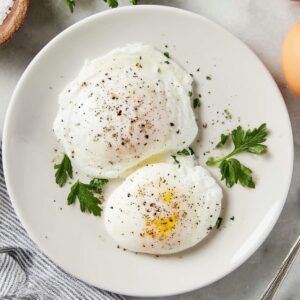 The Best Poached Eggs Recipe 16 Top-down view of two poached eggs on a white plate, lightly seasoned with black pepper, garnished with fresh parsley, accompanied by whole eggs, a bowl of salt, and a striped kitchen towel on a white marble countertop