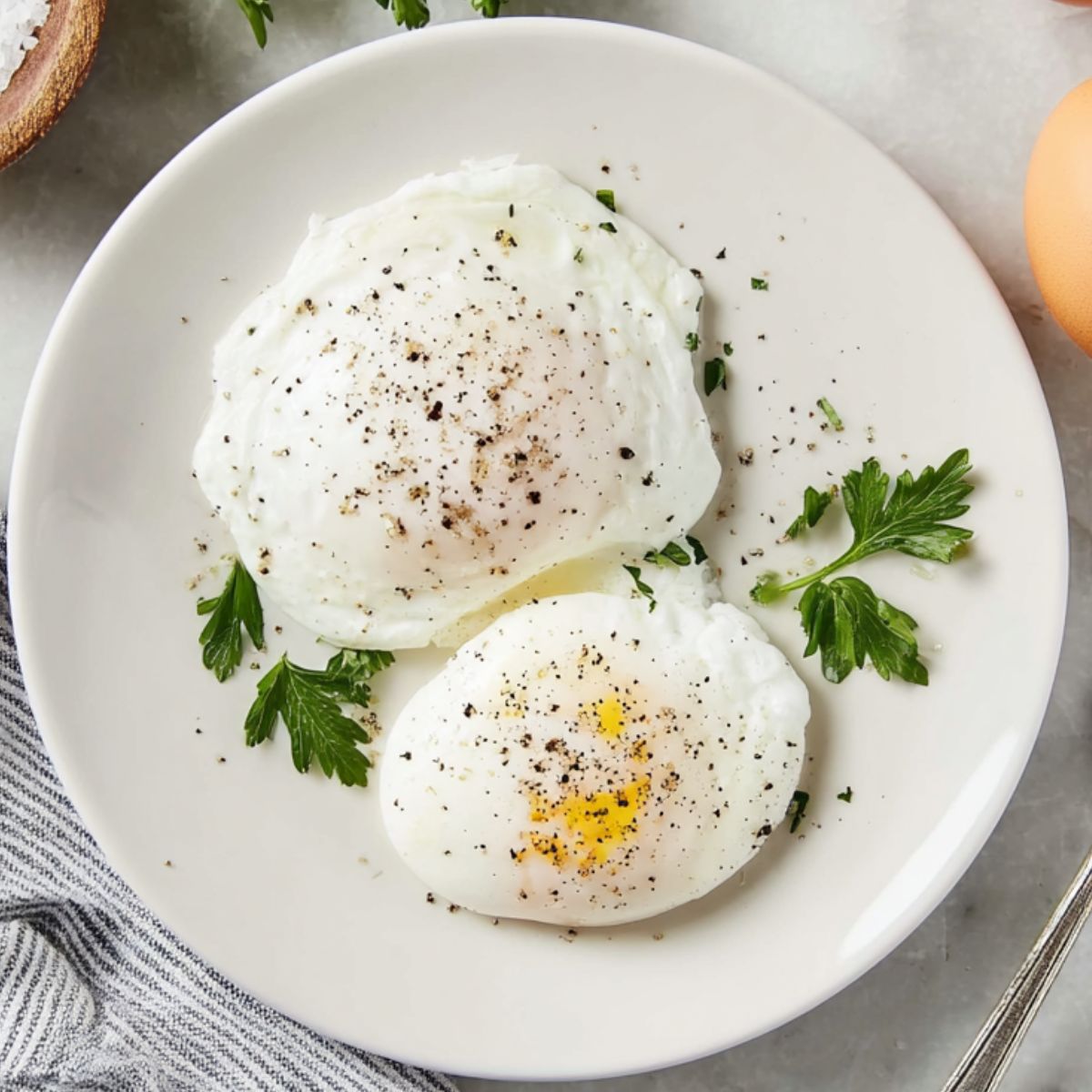 The Best Poached Eggs Recipe 12 Top-down view of two poached eggs on a white plate, lightly seasoned with black pepper, garnished with fresh parsley, accompanied by whole eggs, a bowl of salt, and a striped kitchen towel on a white marble countertop
