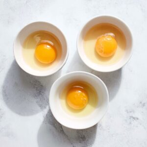 The Best Poached Eggs Recipe 14 Overhead view of three raw eggs cracked into individual white bowls, placed on a white marble countertop, bright natural lighting, focus on vibrant yolks and translucent egg whites.