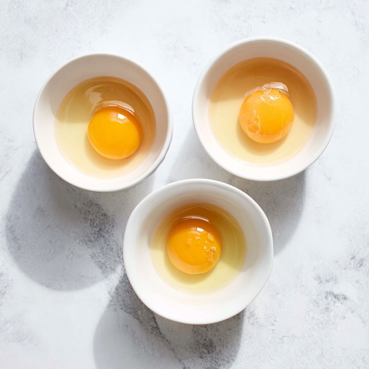 The Best Poached Eggs Recipe 10 Overhead view of three raw eggs cracked into individual white bowls, placed on a white marble countertop, bright natural lighting, focus on vibrant yolks and translucent egg whites.
