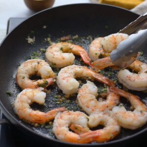 Delicious Prawn And Chorizo Pasta Recipe 16 Raw shrimp arranged in a black pan being cooked, some already turning pink, with tongs lifting a few.