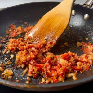 Delicious Prawn And Chorizo Pasta Recipe 17 A close-up of finely chopped shallots and tomato sautéing in a black pan with a wooden spatula.