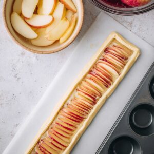 Delicious Puff Pastry Apple Roses Recipe 17 A strip of puff pastry brushed with fruit jam and lined with thin apple slices, ready to be rolled into an apple rose, next to a bowl of apple slices and a muffin tin.