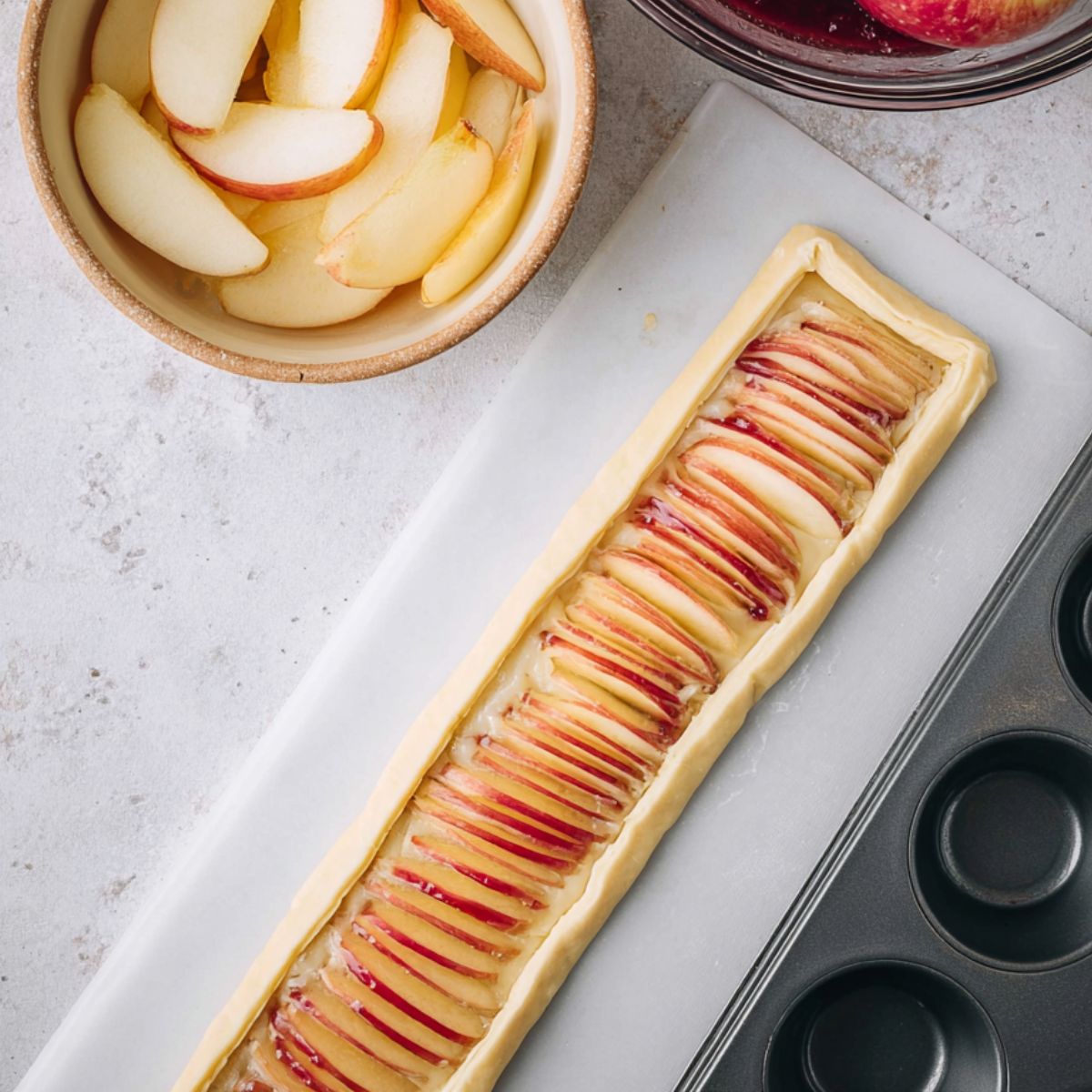 Delicious Puff Pastry Apple Roses Recipe 12 A strip of puff pastry brushed with fruit jam and lined with thin apple slices, ready to be rolled into an apple rose, next to a bowl of apple slices and a muffin tin.