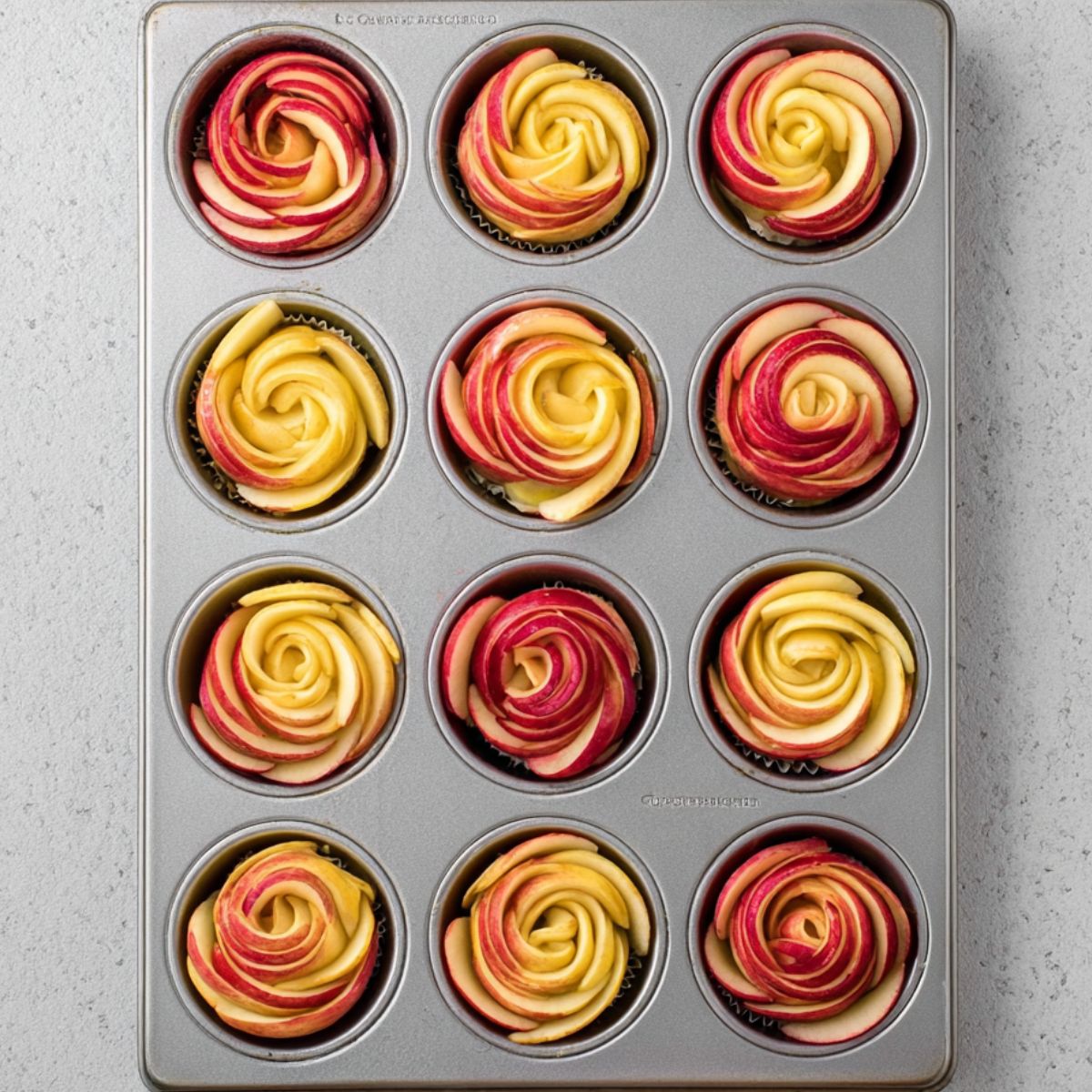 Delicious Puff Pastry Apple Roses Recipe 13 A 12-cup muffin tin filled with apple rose pastries, some made with red apple slices and some with yellow apple slices, arranged neatly before baking.