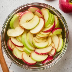 Delicious Puff Pastry Apple Roses Recipe 15 A bowl of thinly sliced red and green apples soaking in water to prevent browning, with two whole apples and a wooden spoon nearby.