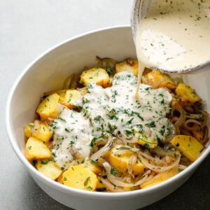 Best Squash Casserole Recipe 17 A bowl of cream and herbs being poured over sautéed squash and onions, ready to be mixed in for a creamy texture and flavorful taste.