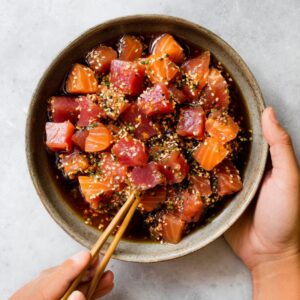 Delicious Sushi Poke Bowls Recipe 14 A top-down view of a bowl filled with cubed raw salmon and tuna, tossed in a soy-based sauce with sesame seeds, being held by one hand while the other hand uses chopsticks to pick up pieces.
