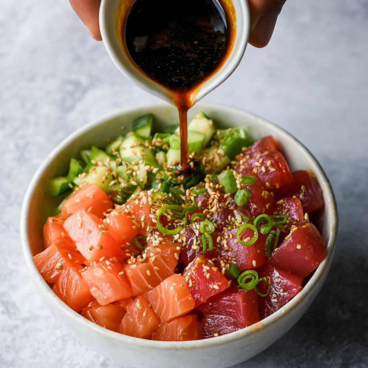 Delicious Sushi Poke Bowls Recipe 10 A poke bowl with cubed raw salmon and tuna, chopped green onions, and sesame seeds, with a small cup of dark soy-based sauce being poured over the bowl.