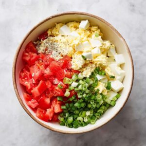 Healthy Tomato Cracker Salad Recipe 15 A bowl filled with diced tomatoes, chopped boiled eggs, green onions, and mayonnaise, seasoned with salt and pepper, ready to be mixed into a fresh salad.