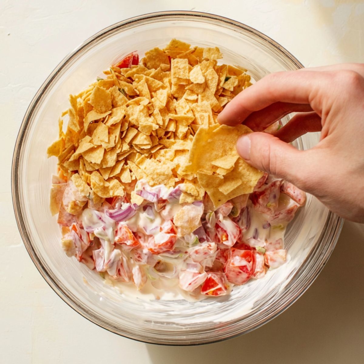 Healthy Tomato Cracker Salad Recipe 12 A hand placing crushed crackers into a creamy mixture of tomatoes, onions, and dressing in a bowl, likely preparing a refreshing salad or snack