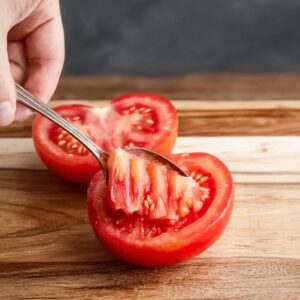 Healthy Tomato Cracker Salad Recipe 14 A hand carefully deseeding a ripe tomato using a spoon, with the tomato halves placed on a wooden cutting board, preparing for a recipe