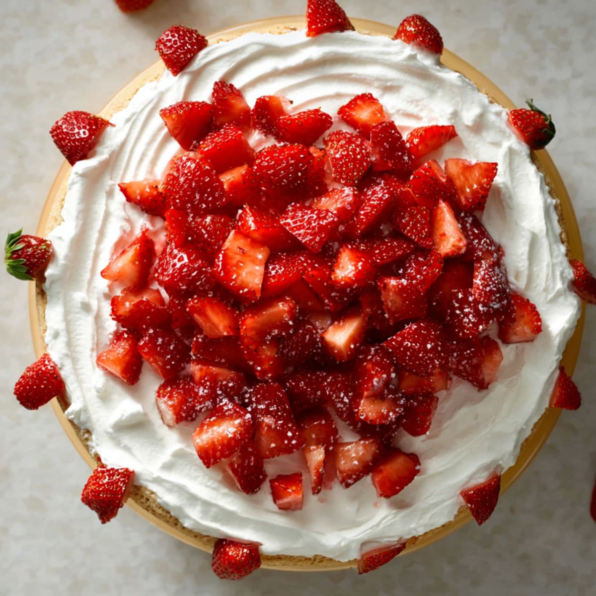 The Best Vegan Strawberry Shortcake Cake 13 A top-down view of a strawberry shortcake with a smooth layer of whipped cream and fresh chopped strawberries scattered on top, garnished with whole strawberries along the edges.
