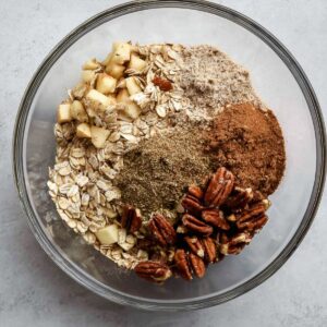 A glass bowl with dry ingredients for baked apple oatmeal, including oats, chopped pecans, cinnamon, and ground flaxseed.