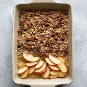 A rectangular dish of baked apple oatmeal showing a layer of cooked apple slices underneath an oat and pecan topping, partially spread before baking.