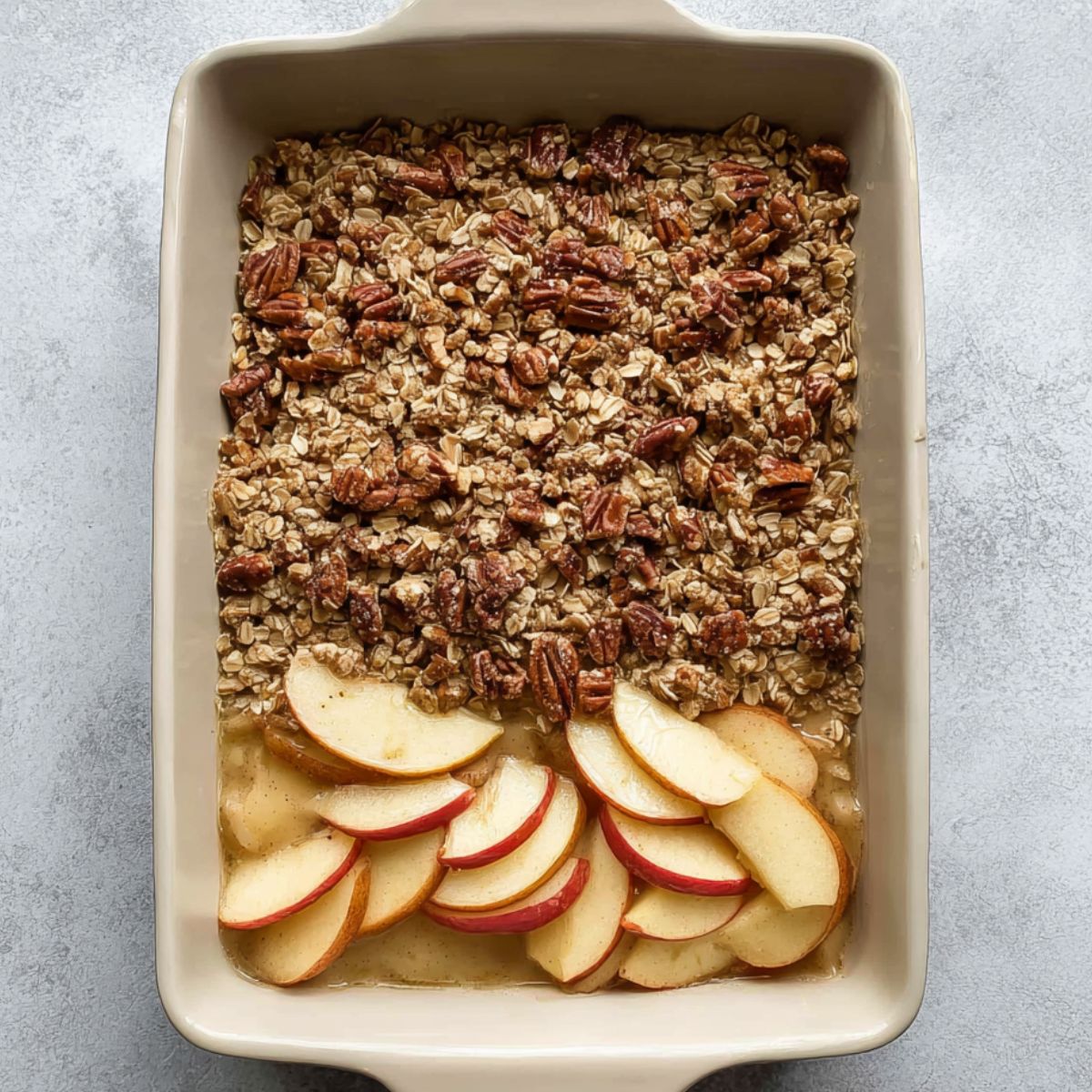 Best Baked Apple Oatmeal Recipe 12 A rectangular dish of baked apple oatmeal showing a layer of cooked apple slices underneath an oat and pecan topping, partially spread before baking.