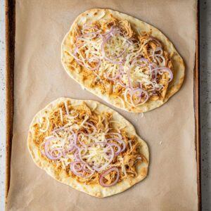 Best Buffalo Chicken Flatbreads Recipe 15 "Two flatbreads on a parchment-lined baking sheet, topped with shredded chicken, red onion slices, and shredded mozzarella, ready to bake.