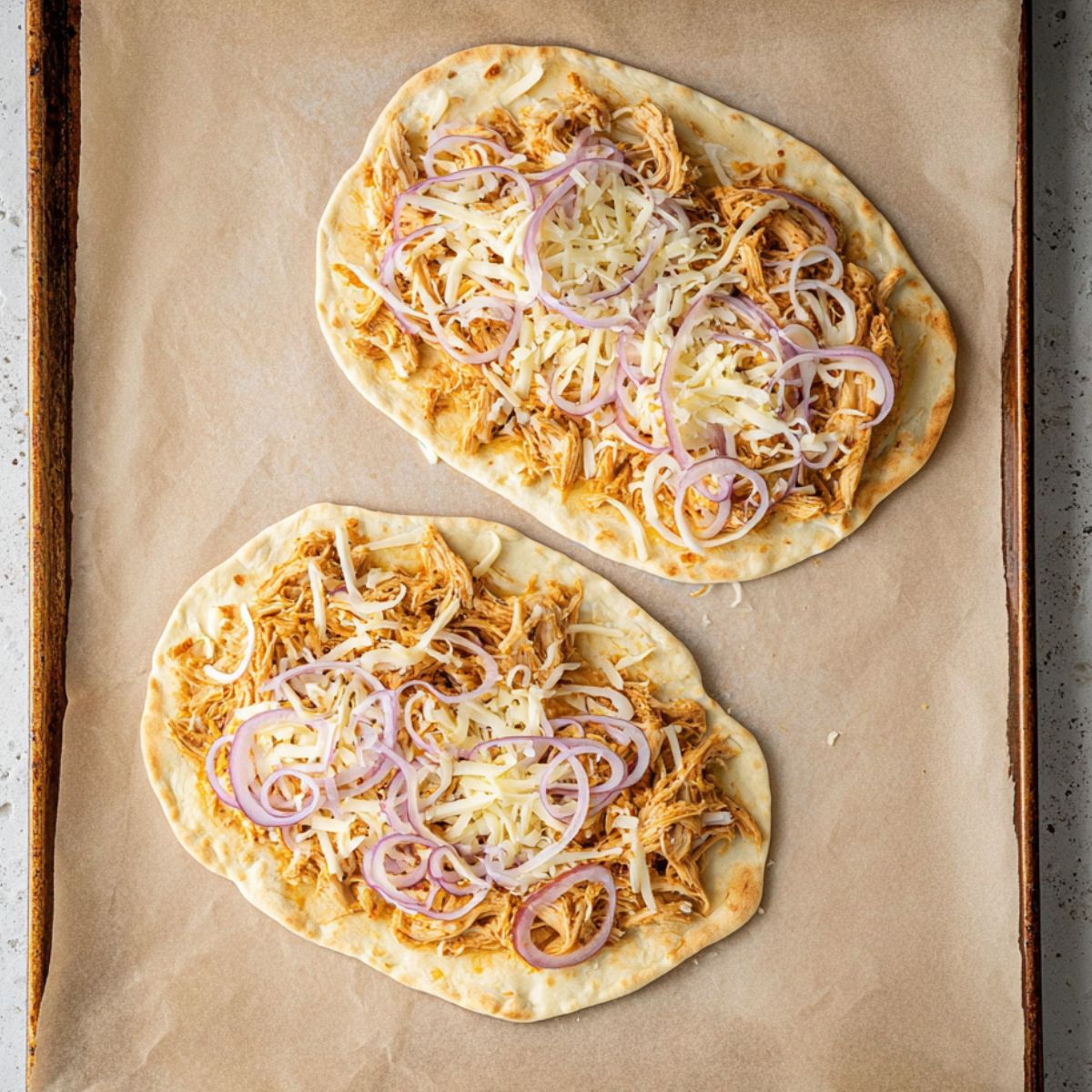 Best Buffalo Chicken Flatbreads Recipe 11 "Two flatbreads on a parchment-lined baking sheet, topped with shredded chicken, red onion slices, and shredded mozzarella, ready to bake.