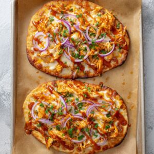 Best Buffalo Chicken Flatbreads Recipe 16 Two baked buffalo chicken flatbreads on a parchment-lined baking sheet, topped with shredded chicken, melted cheese, red onion slices, and drizzled with buffalo sauce.