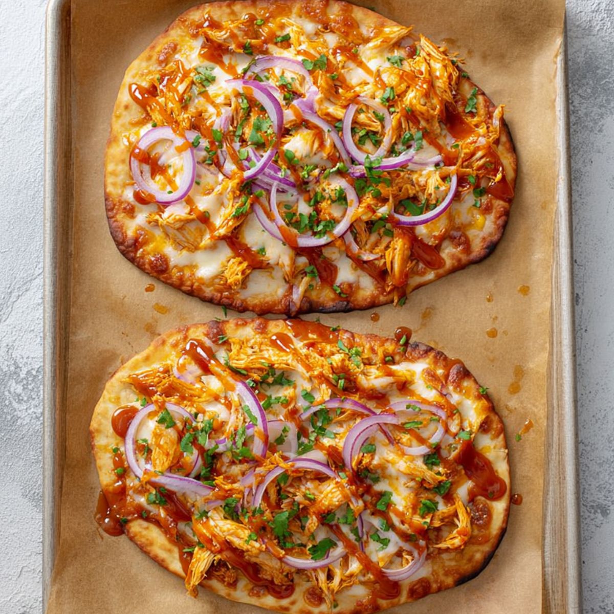 Best Buffalo Chicken Flatbreads Recipe 12 Two baked buffalo chicken flatbreads on a parchment-lined baking sheet, topped with shredded chicken, melted cheese, red onion slices, and drizzled with buffalo sauce.