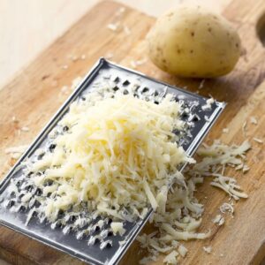 Delicious Crispy Hash Browns Recipe 14 A peeled potato on a metal grater next to a pile of freshly shredded potato, placed on a wooden cutting board, top-down view, bright natural lighting, kitchen food preparation scene, high-resolution, clean and minimalistic composition, focus on texture and detail of potato shreds.
